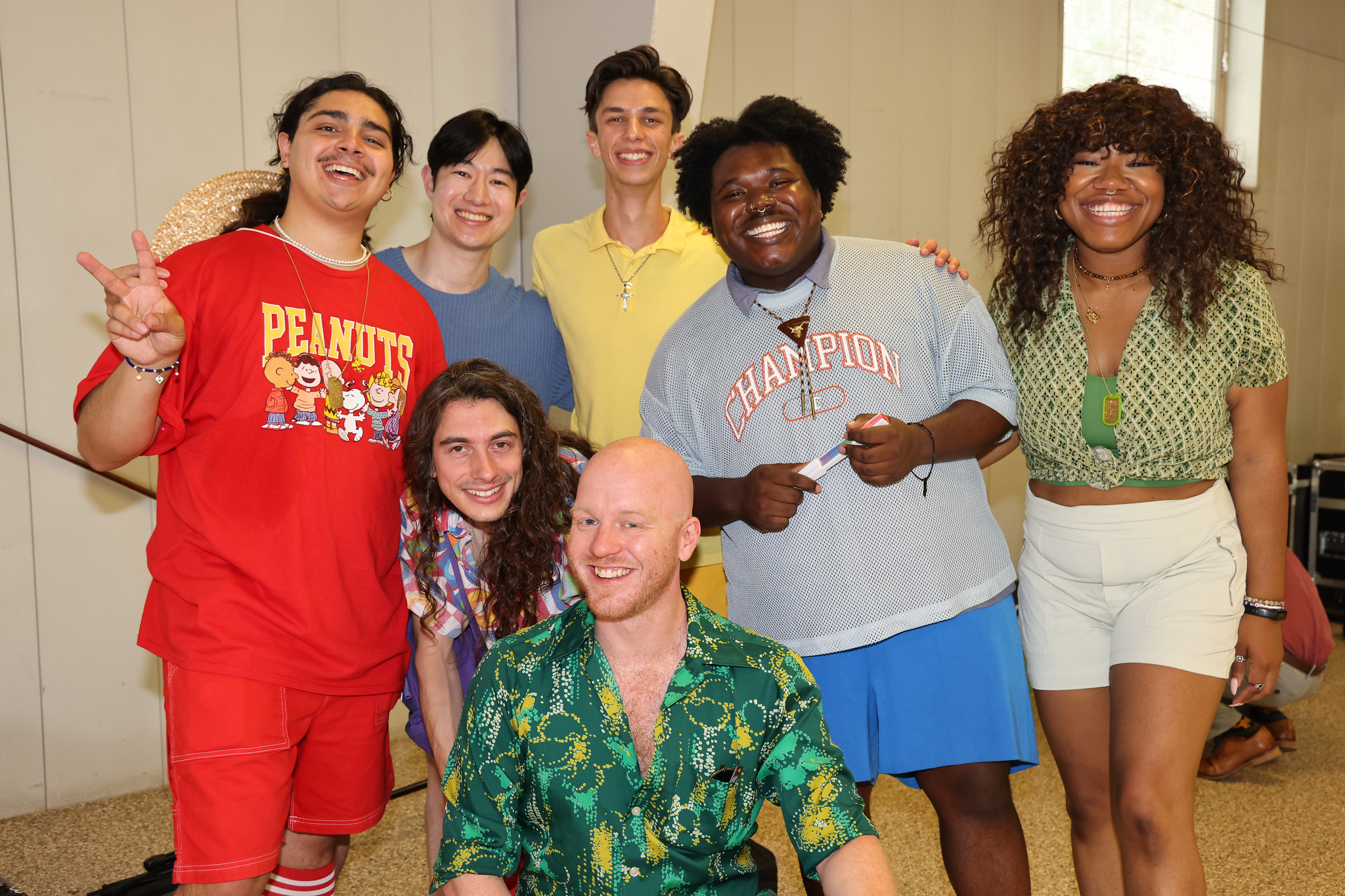 Group of seven diverse people smiling, one wearing a red "Peanuts" shirt, gathered in casual attire indoors