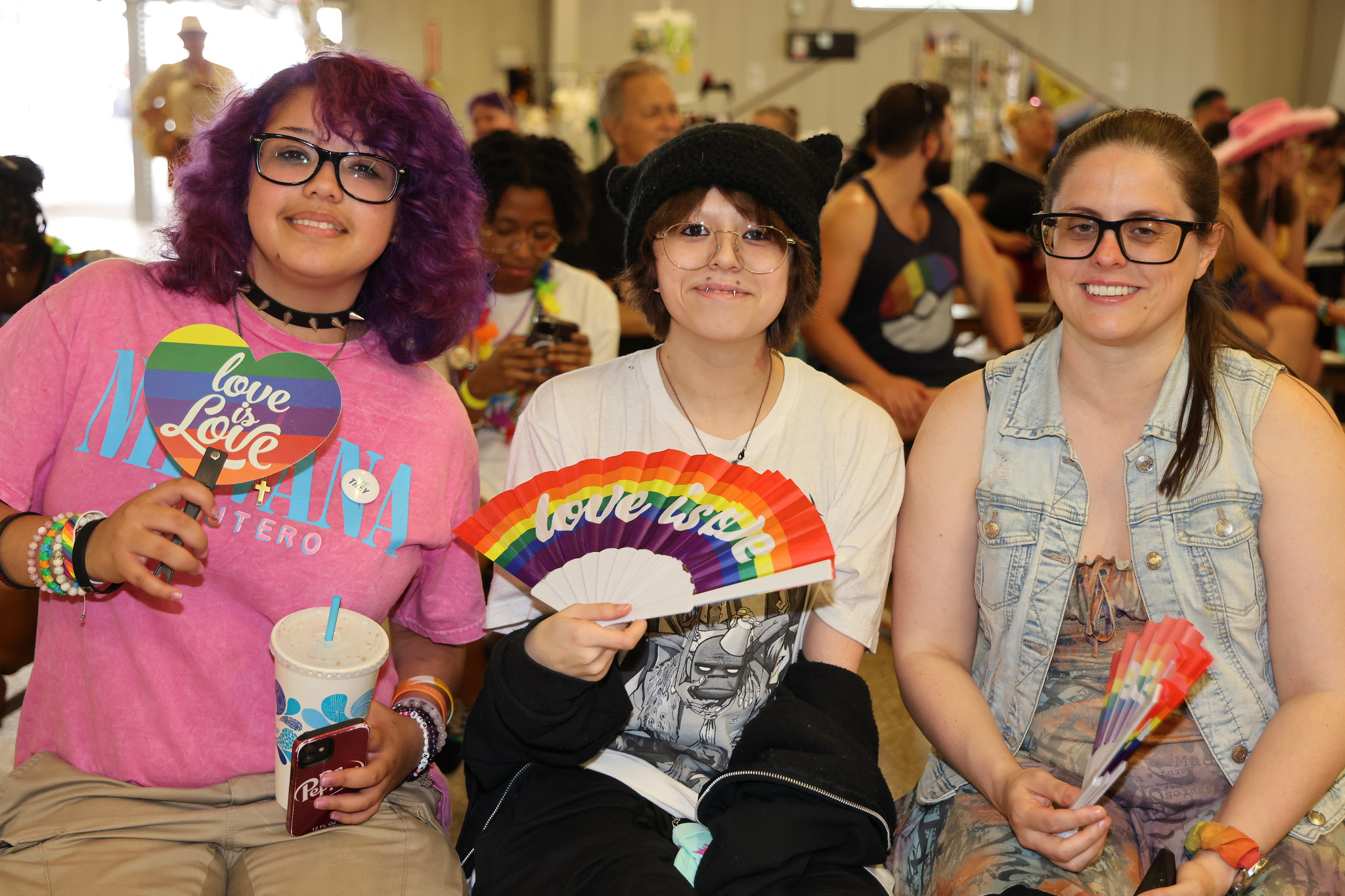Three people smiling, holding rainbow fans, and wearing "love is love" items in a festive gathering