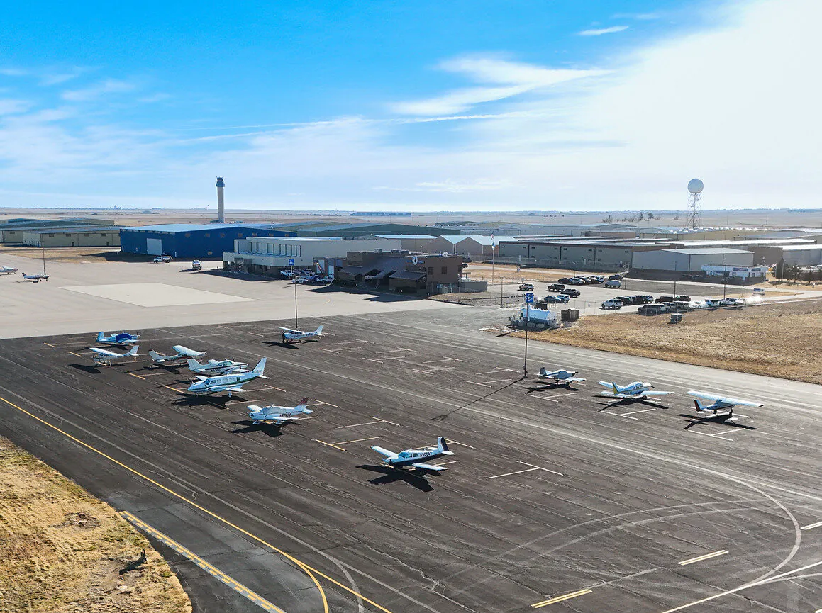 Several small airplanes parked on an airport tarmac with buildings nearby
