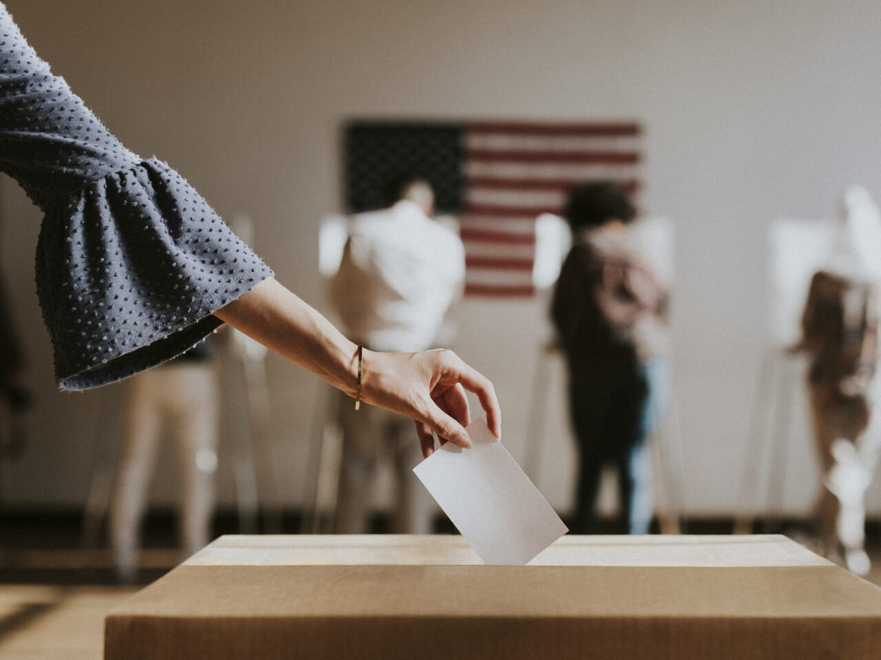Hand dropping a ballot into a box with voters and a U.S. flag blurred in the background
