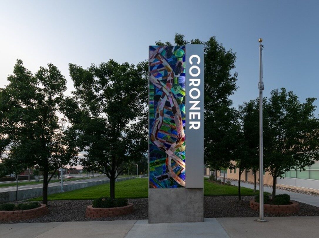 Tall sign labeled "CORONER" with abstract mosaic beside trees and flagpole