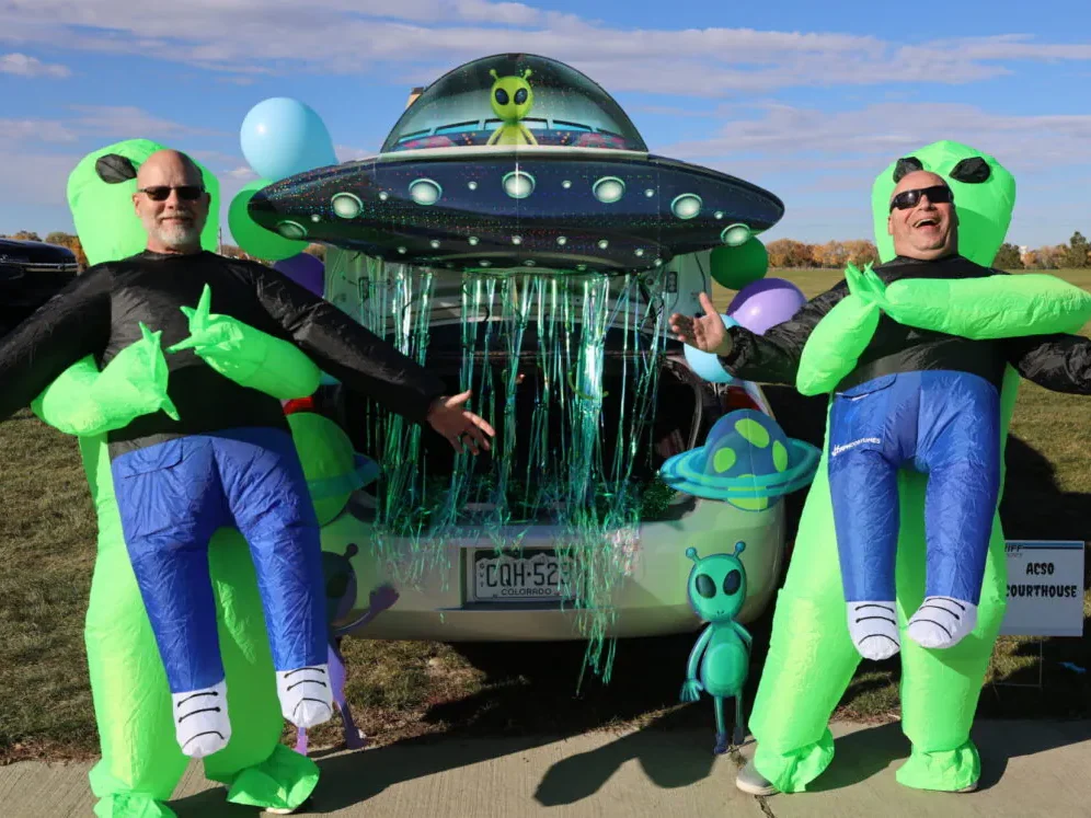 Two people in inflatable alien costumes pose joyfully beside a UFO-themed car trunk display with colorful decorations