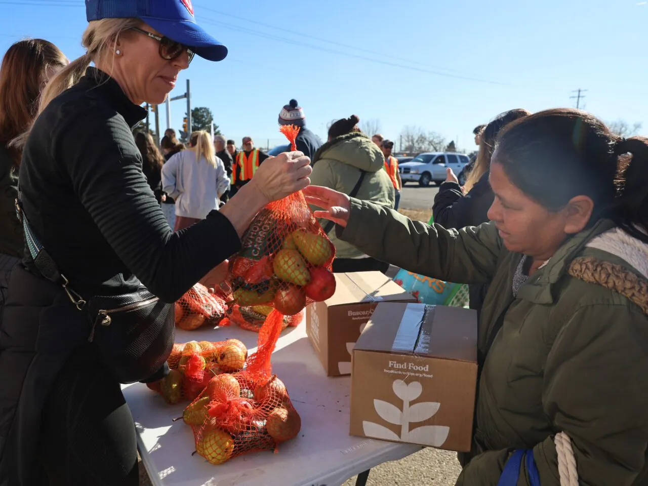 Woman in cap distributing bags of produce to another woman at outdoor food drive. Cardboard boxes labeled "Find Food."