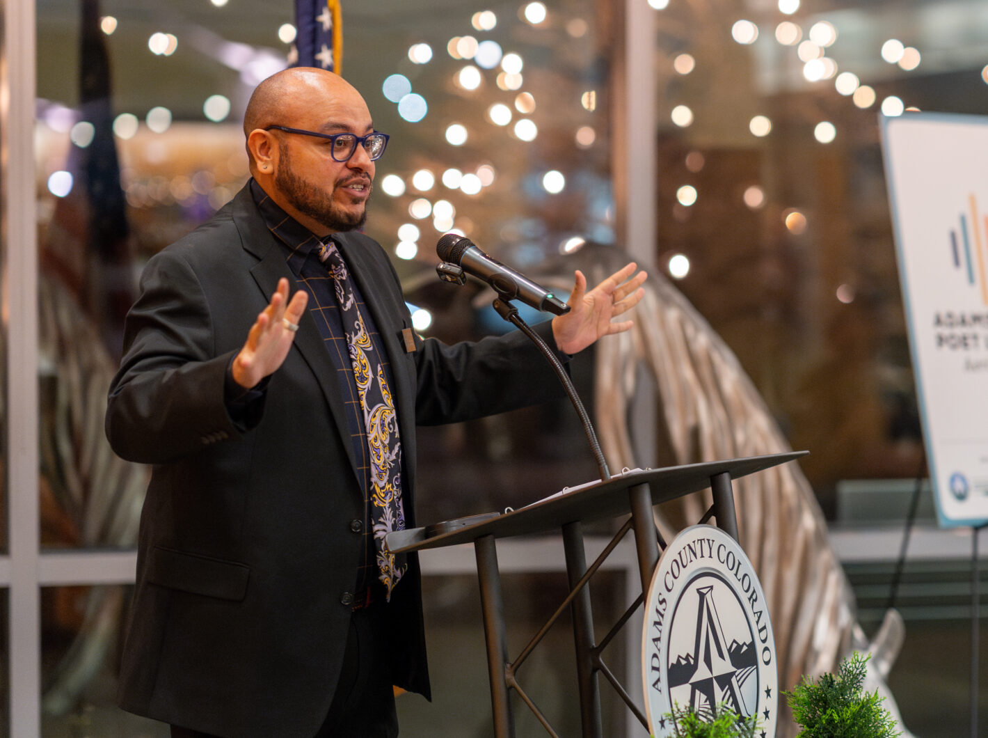 Poet Laureate speaks energetically at a podium during an event, with sparkling lights in the background