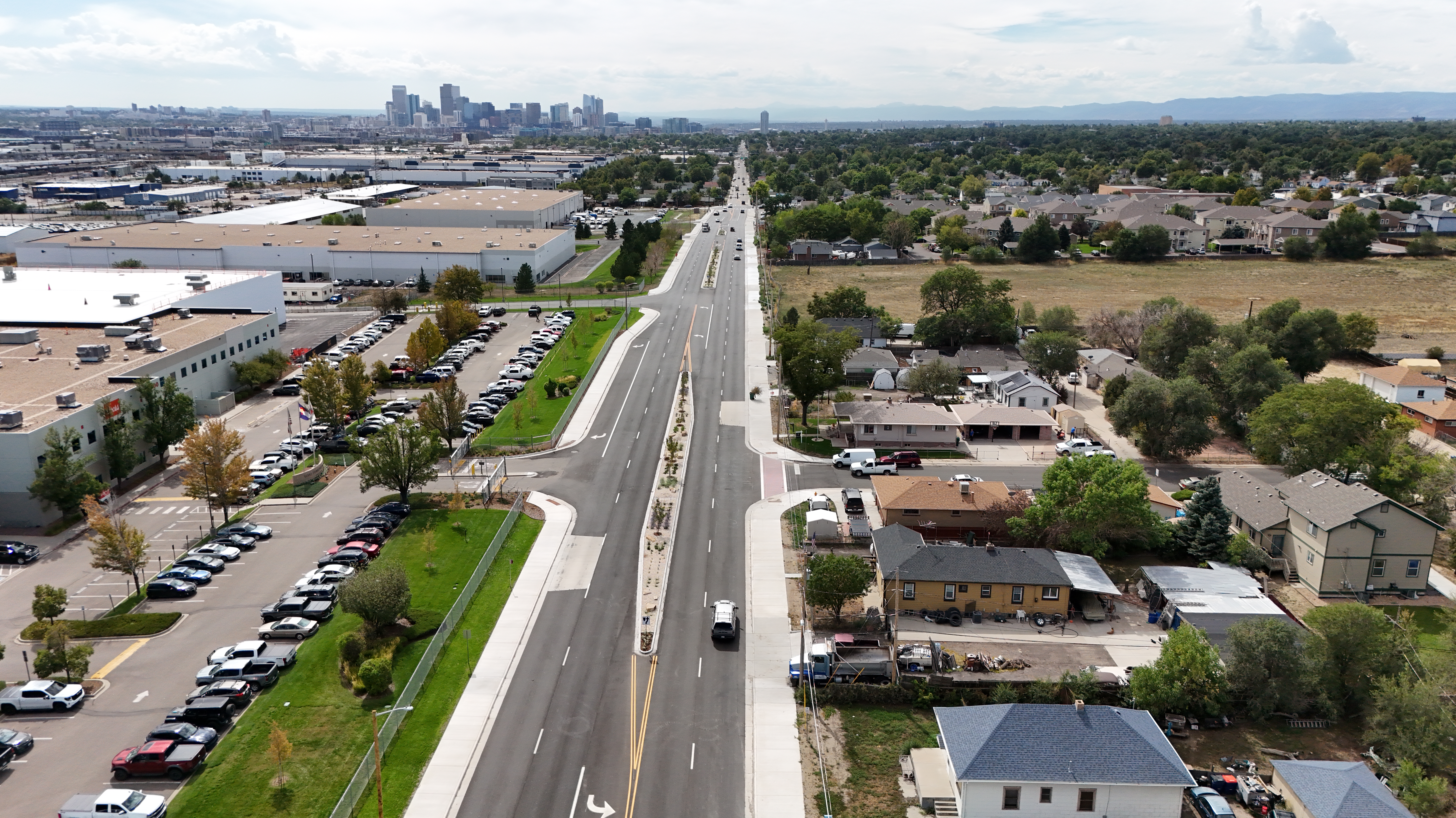 Aerial view of a wide road dividing industrial buildings and residential area, leading towards a distant city skyline