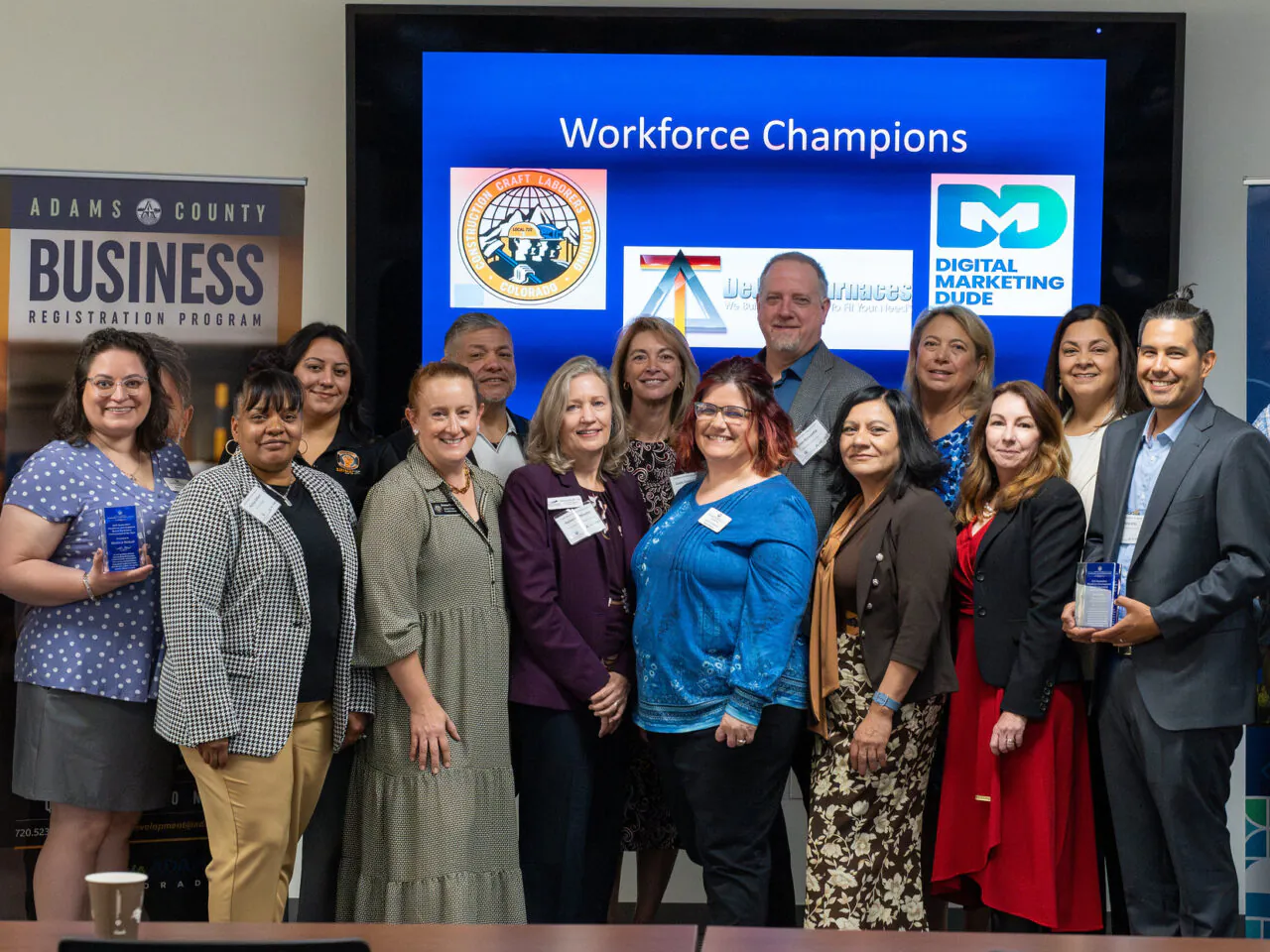 Group of diverse professionals posing with awards, under "Workforce Champions" display, part of Adams County Business event