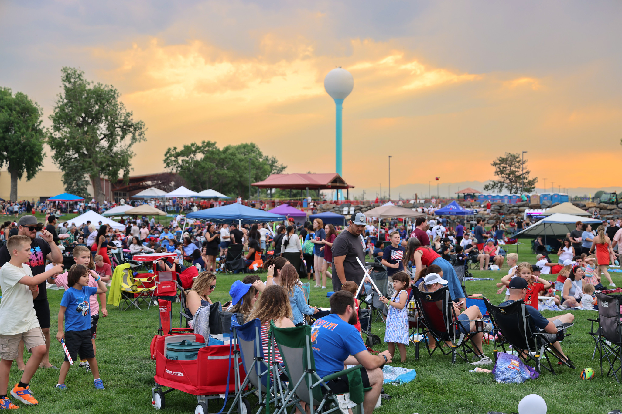 Crowd enjoying an outdoor event at sunset in a park, with families, children playing, tents, and a water tower in the background