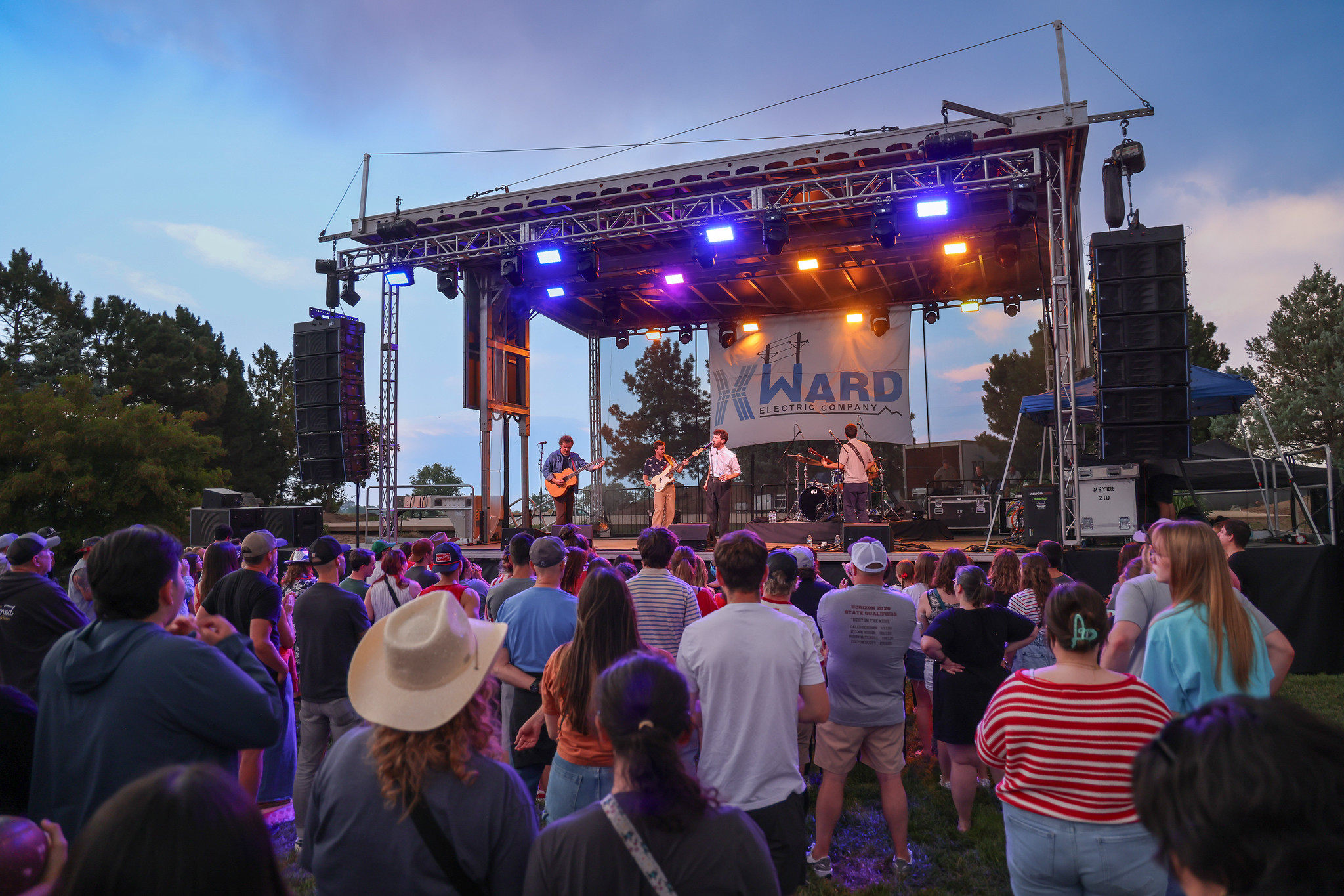 Outdoor concert with a crowd watching a band on stage under colorful lights, Ward Electric Company banner displayed above