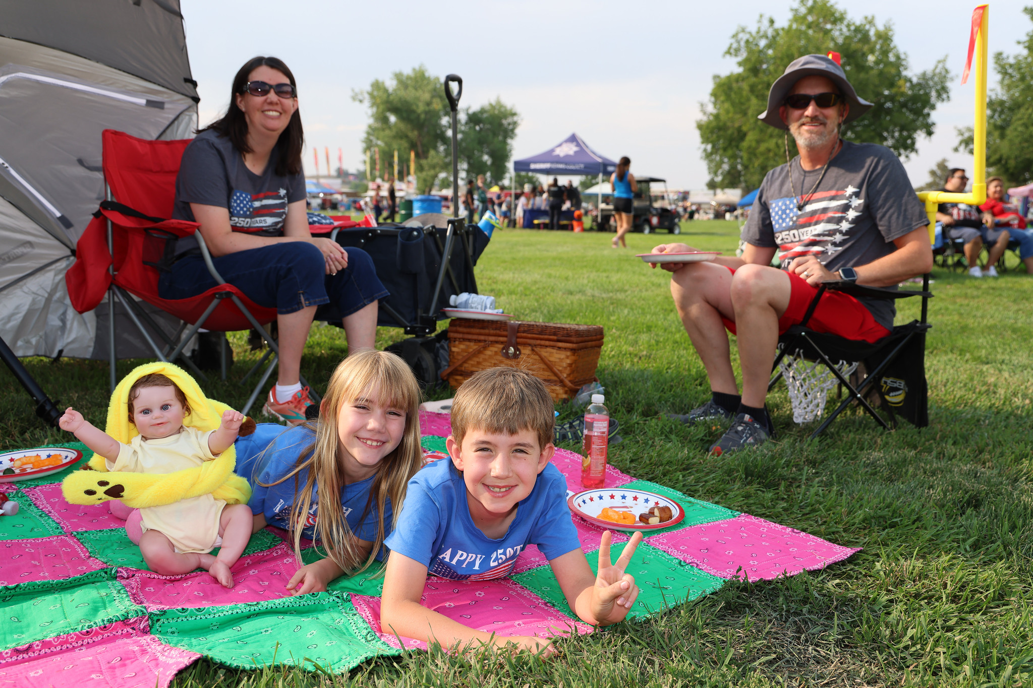 Family with young children on a picnic blanket, enjoying a sunny day outdoors. Adults wear shirts with American flag designs