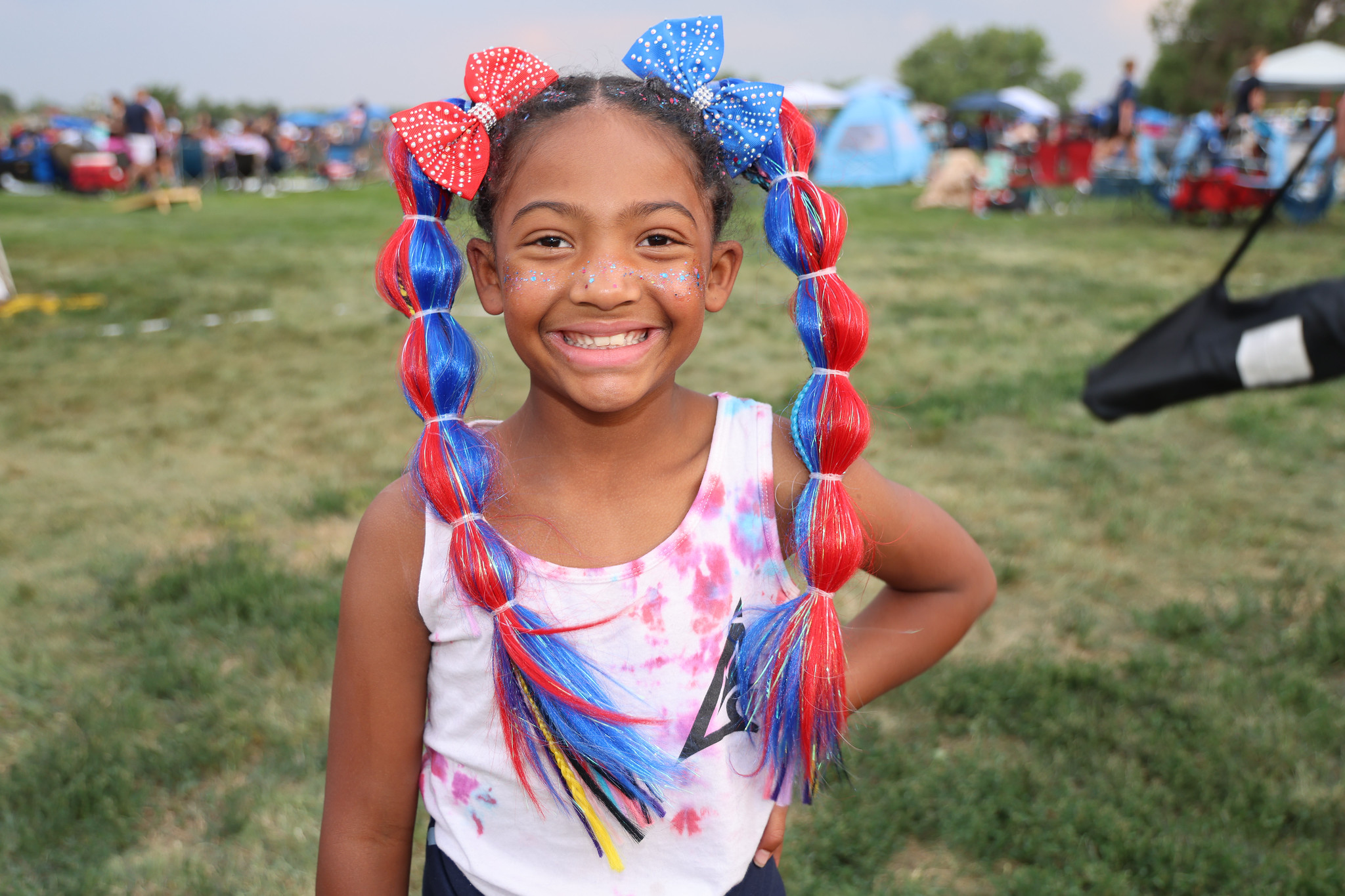 Young girl smiling brightly with red, white, and blue braided hair, wearing a tie-dye shirt, outdoors at a festive event