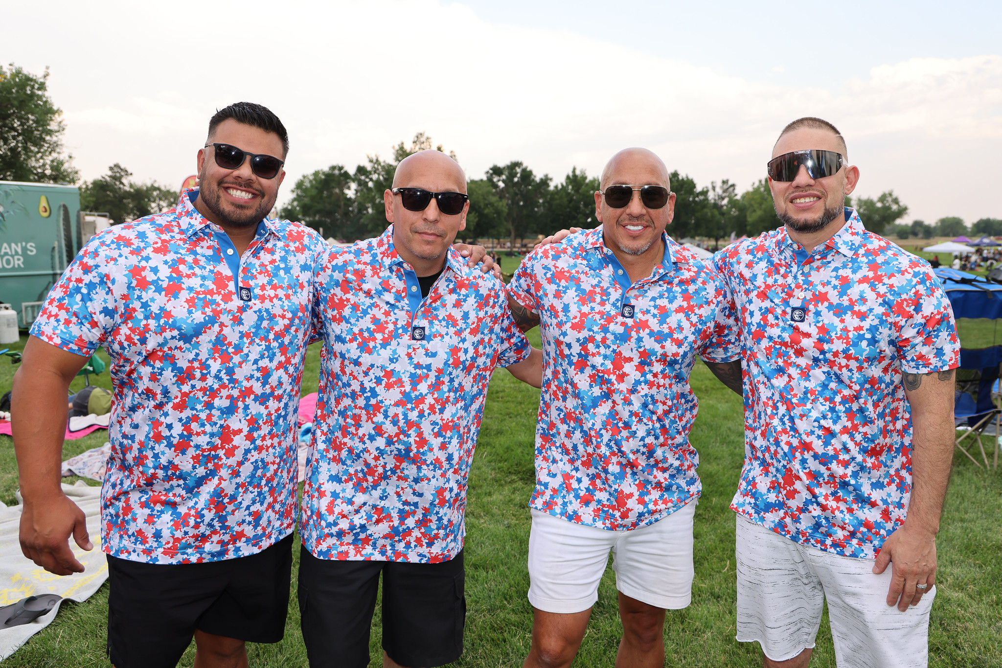 Four men in matching red, white, and blue star-patterned shirts, smiling and wearing sunglasses at a park event