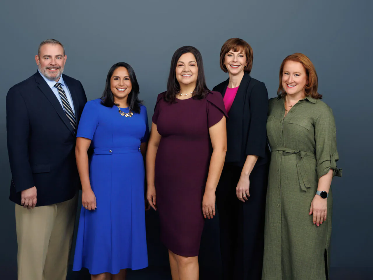 Five people standing together, smiling, in professional attire, against a plain gray background