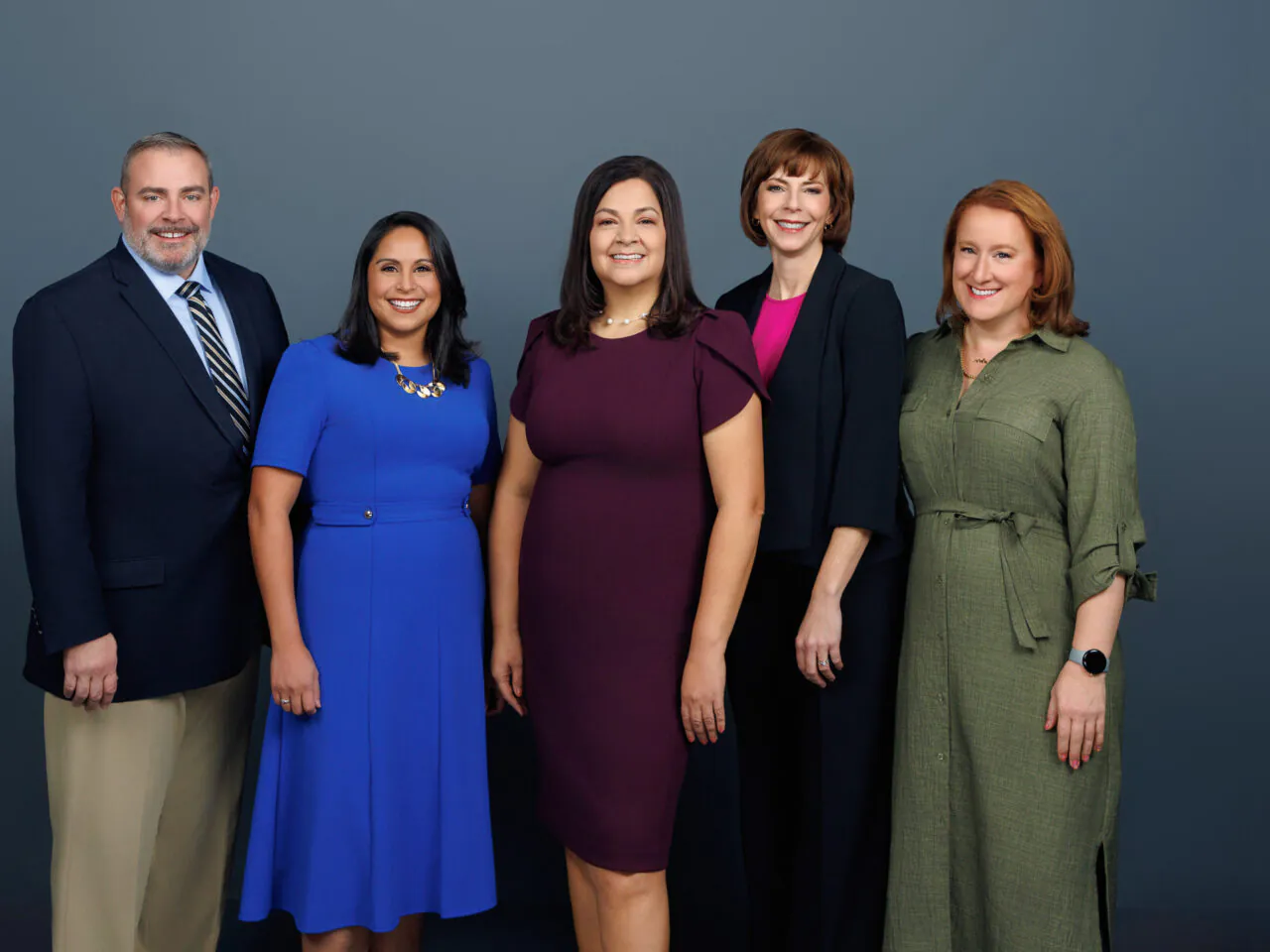 Five professionals smiling against a plain background, dressed in business attire, suggesting a formal group portrait