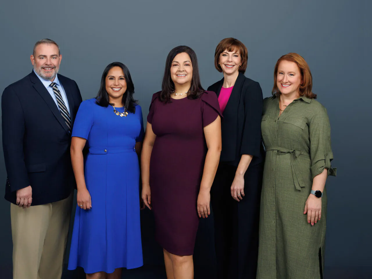 Five people standing together, smiling, against a plain gray background