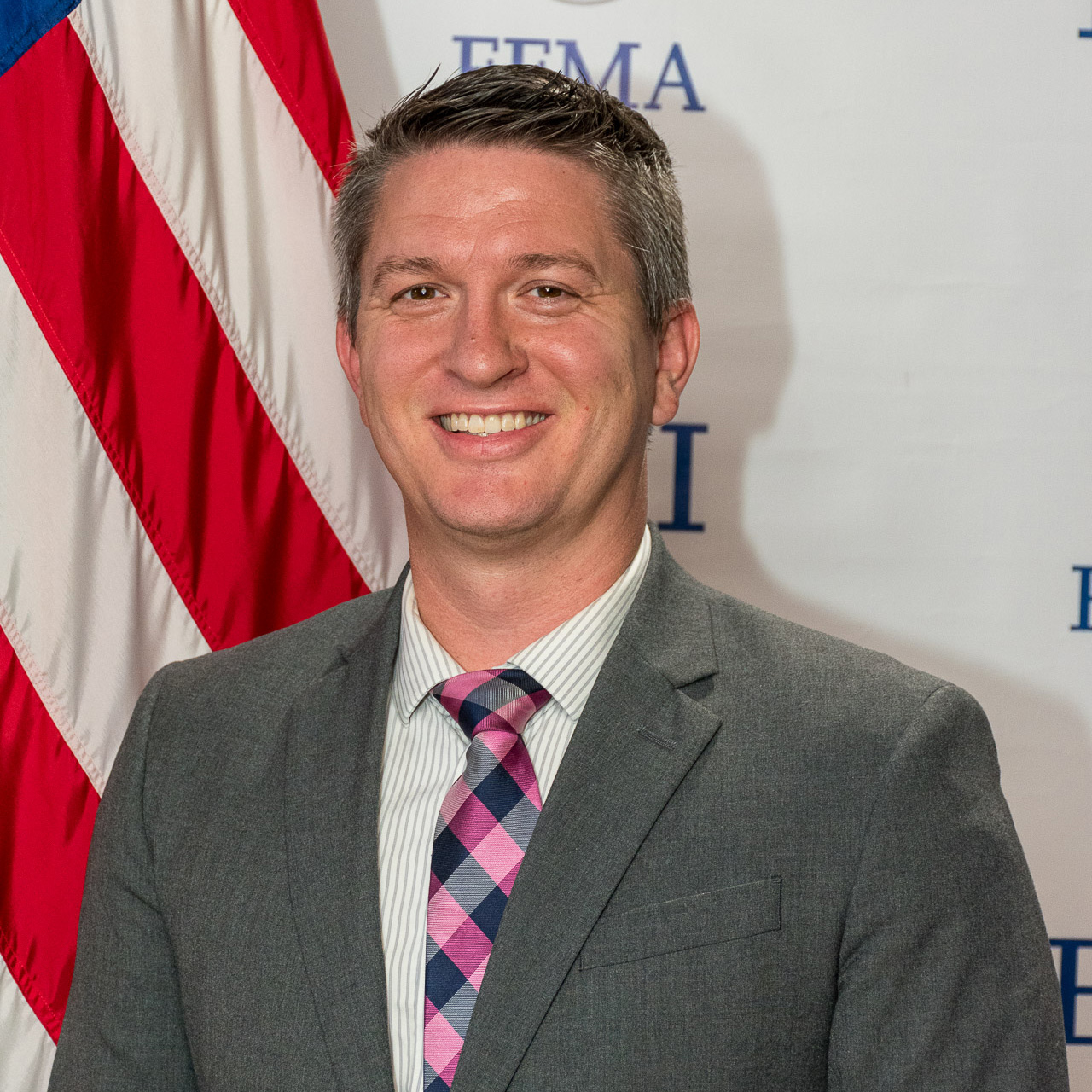 Man in a suit and patterned tie smiling, standing in front of an American flag and a FEMA backdrop