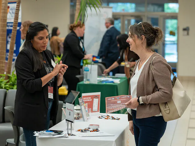 Two women engaged in conversation at an event booth with informational materials and small items on the table