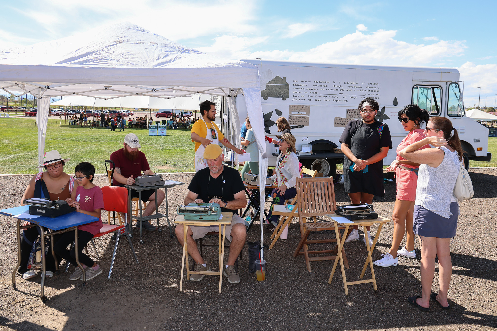 Community event with people at typewriters under a tent, next to an arts truck in a sunny outdoor setting