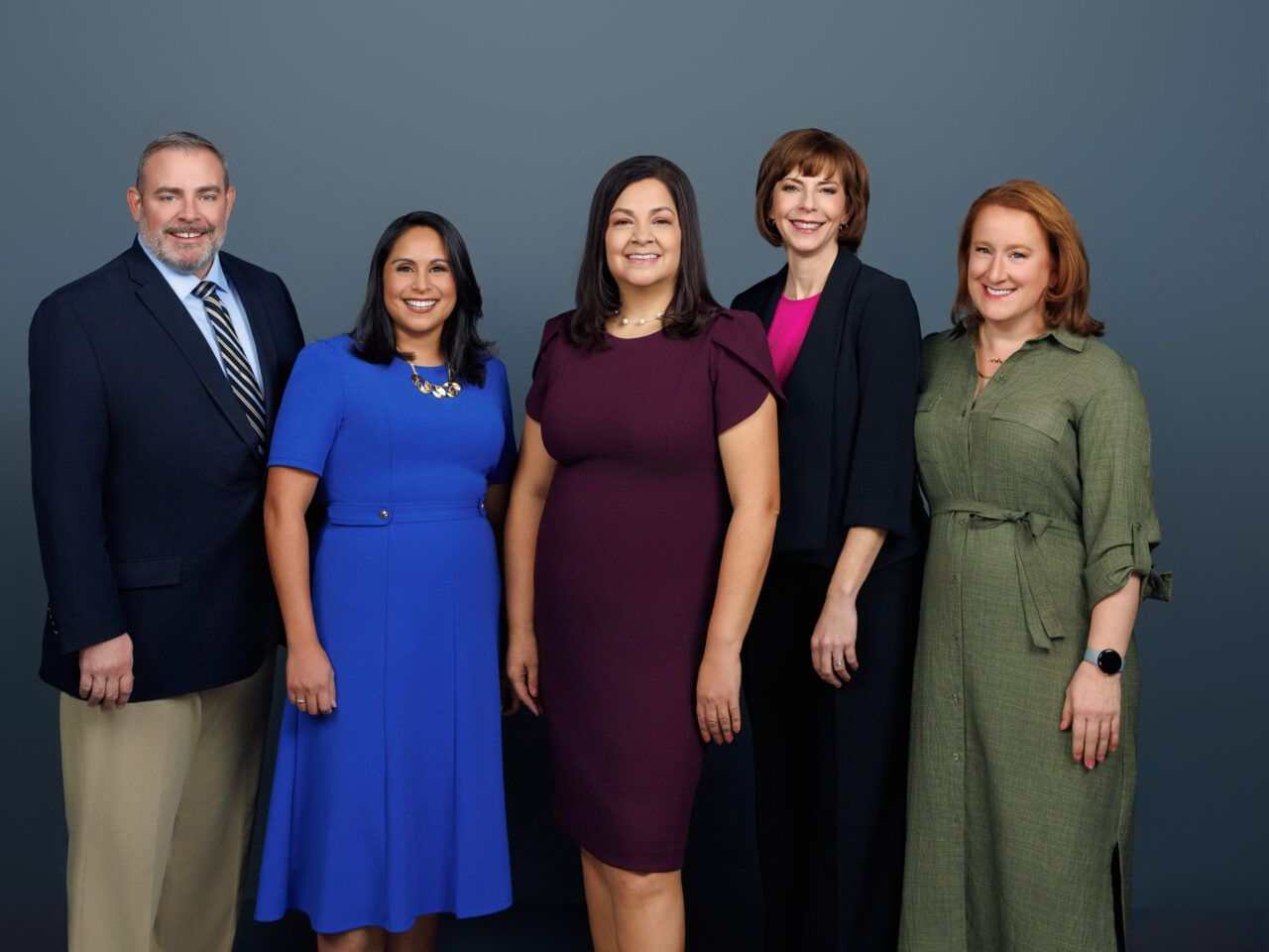 Five people standing side by side in formal attire against a neutral backdrop, smiling at the camera