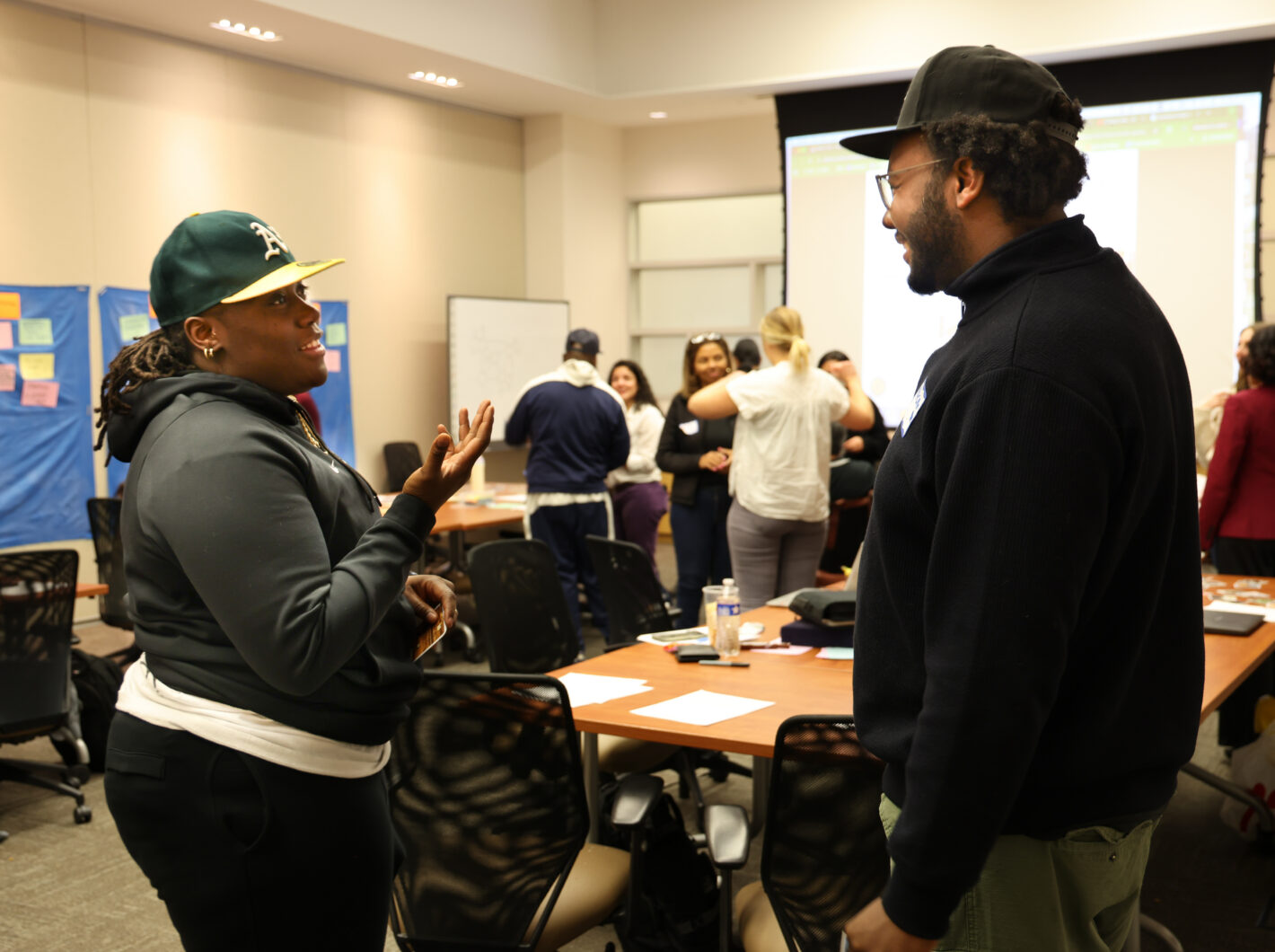 Two people in casual attire engaged in conversation at a community planning meeting, with others in background