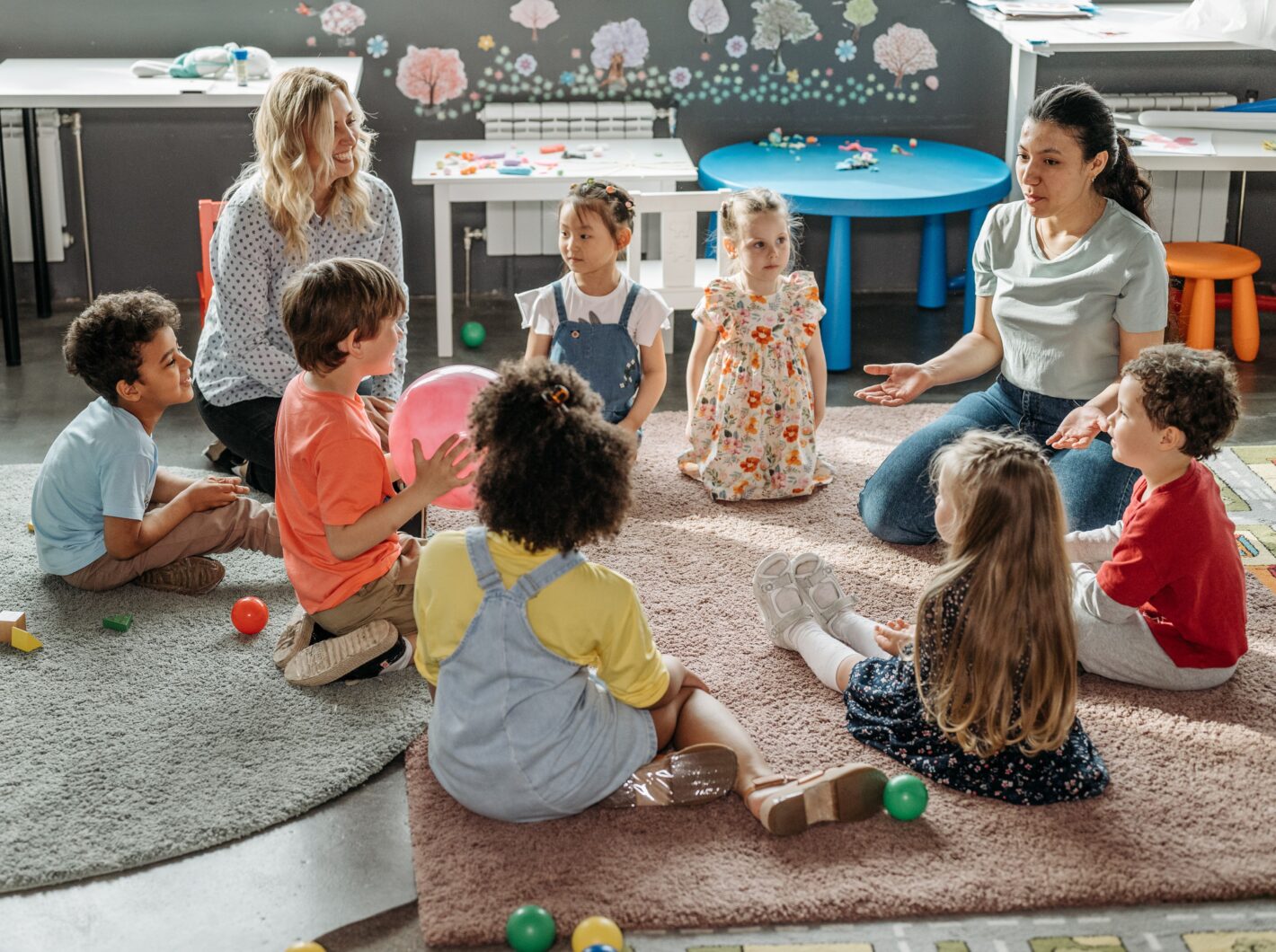 Children and two teachers sit in a circle on the carpet during a classroom activity with colorful toys around them