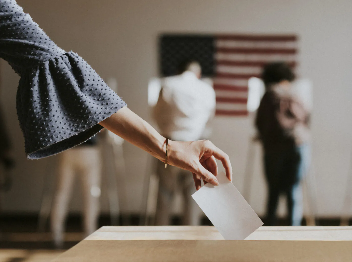 Hand dropping a ballot into a box with voters and a U.S. flag blurred in the background